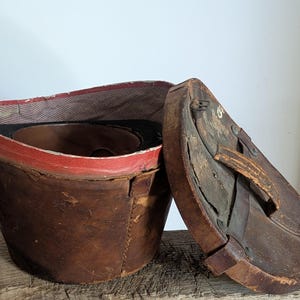 Pu&ograve; includere: Una cappelliera vintage in pelle marrone con coperchio rimovibile. Il contenitore cilindrico presenta una fodera interna in tessuto rosso e una superficie esterna usurata. Il coperchio include una maniglia in pelle e fissaggi in metallo.