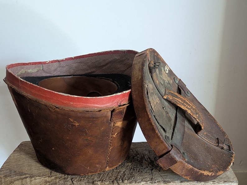 Pu&ograve; includere: Una cappelliera vintage in pelle marrone con coperchio rimovibile. Il contenitore cilindrico presenta una fodera interna in tessuto rosso e una superficie esterna usurata. Il coperchio include una maniglia in pelle e fissaggi in metallo.