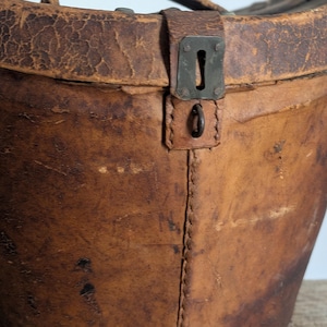 Pu&ograve; includere: Un astuccio cilindrico in pelle marrone usurata con superficie testurizzata, una cucitura verticale e un meccanismo di chiusura in metallo con foro per chiave. L'astuccio include un manico in pelle fissato al coperchio con rivetti in metallo.