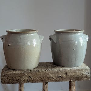 May include: Two ceramic crocks with side handles and a glazed finish. The left crock is a light beige color, while the right crock is a cool grey tone. Both vessels feature a wide mouth, a thick rim, and horizontal ridges near the top opening.