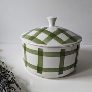 May include: White ceramic lidded jar with a green plaid pattern. The jar has a rounded shape with a small, white knob on the lid. The green plaid design covers the lid and the body of the jar.