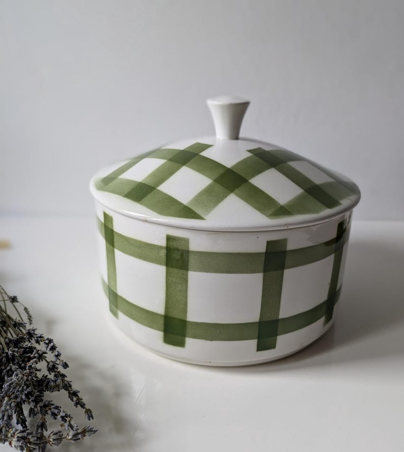 May include: White ceramic lidded jar with a green plaid pattern. The jar has a rounded shape with a small, white knob on the lid. The green plaid design covers the lid and the body of the jar.