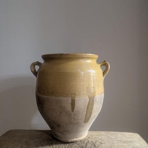 Pode incluir: Um grande recipiente de cerâmica redondo com duas pequenas alças laterais. A parte superior é revestida com um esmalte brilhante amarelo-mostarda que escorre para a parte inferior de argila bege claro, fosca e sem esmalte.