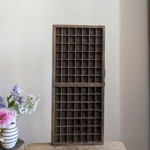 May include: A vintage wooden printer's tray with numerous small compartments. The tray is divided into two sections and has a dark, aged appearance. It sits on a wooden surface, with a vase of flowers to the left.