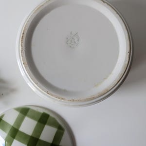 May include: White ceramic serving dish with a lid featuring a green and white checkered pattern. The dish's base is white with a circular design and a faded emblem. The lid is off to the side.