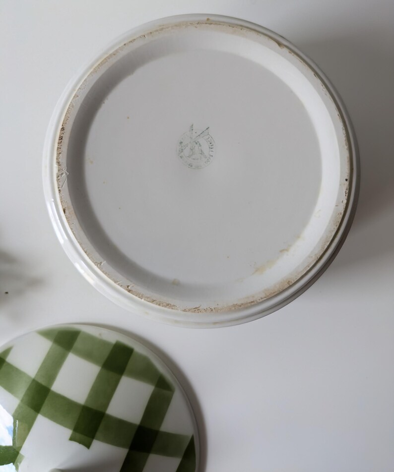 May include: White ceramic serving dish with a lid featuring a green and white checkered pattern. The dish's base is white with a circular design and a faded emblem. The lid is off to the side.