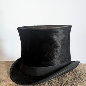 Pu&ograve; includere: Un cappello a cilindro nero con una corona cilindrica alta e una tesa curva. Il cappello presenta una finitura testurizzata simile alla pelliccia sulla corona e un nastro nero liscio e solido che circonda la base dove la corona incontra la tesa.