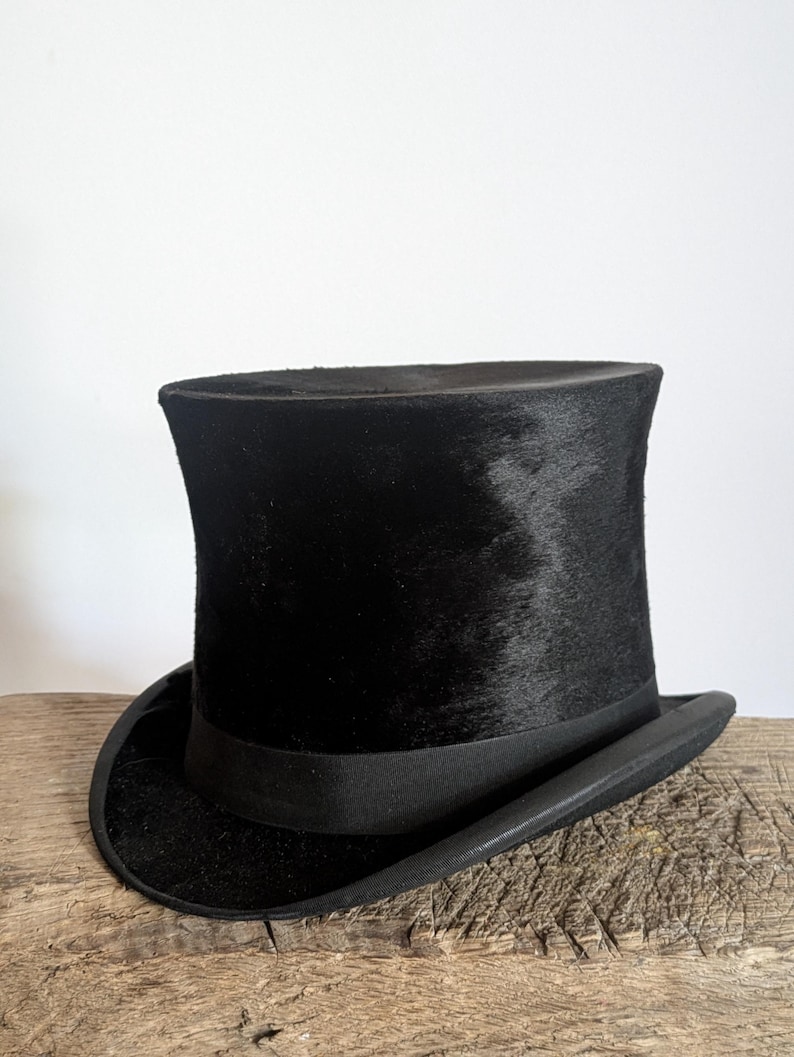 Pu&ograve; includere: Un cappello a cilindro nero con una corona cilindrica alta e una tesa curva. Il cappello presenta una finitura testurizzata simile alla pelliccia sulla corona e un nastro nero liscio e solido che circonda la base dove la corona incontra la tesa.