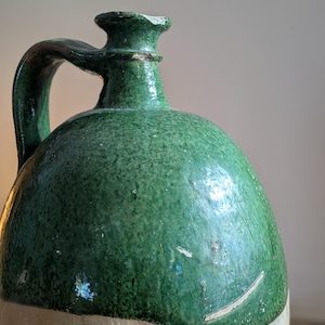 May include: A vintage green glazed stoneware jug with a handle. The jug has a rounded body and a light-colored base. The green glaze shows signs of wear, adding to its antique charm. The jug's design suggests it was used for storing liquids.