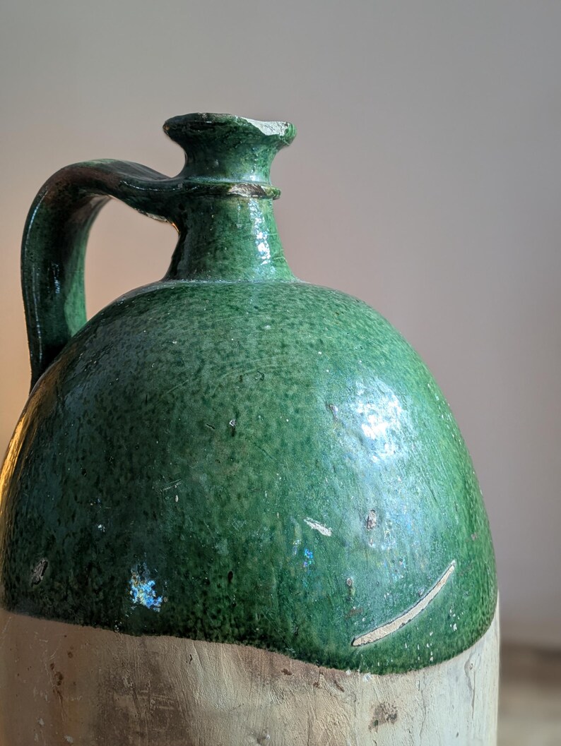 May include: A vintage green glazed stoneware jug with a handle. The jug has a rounded body and a light-colored base. The green glaze shows signs of wear, adding to its antique charm. The jug's design suggests it was used for storing liquids.
