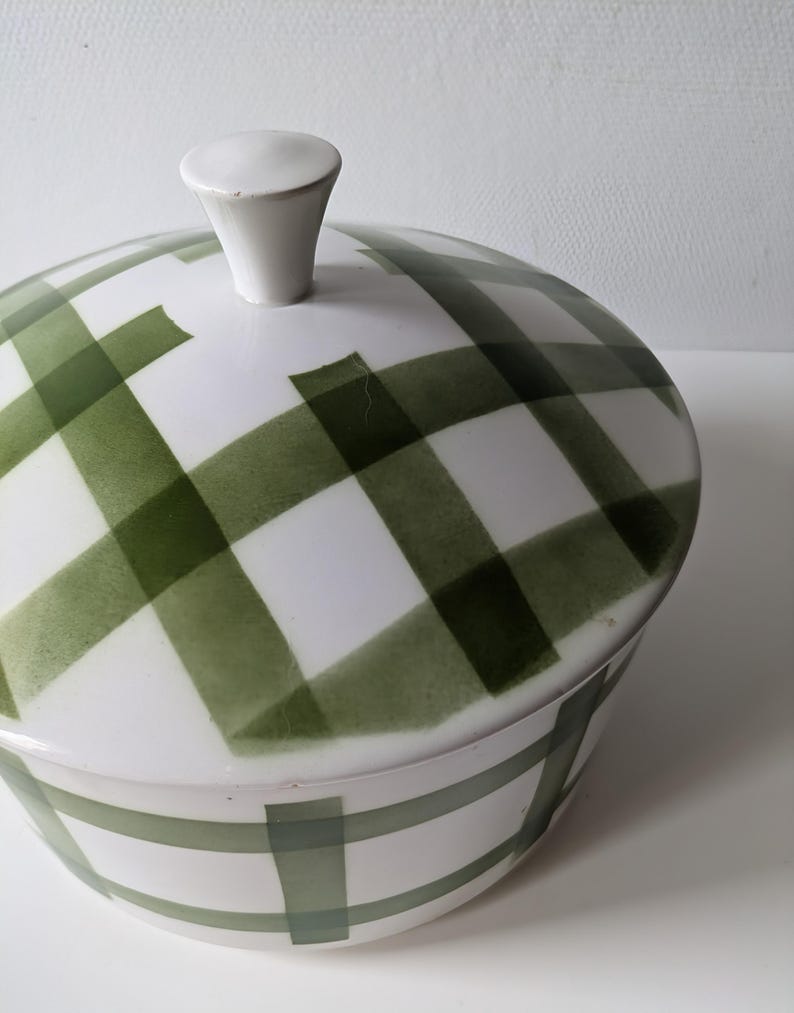 May include: A white ceramic serving dish with a lid, featuring a green plaid pattern. The lid has a small, white knob. The dish is designed for serving food and adds a touch of vintage charm to any table setting.