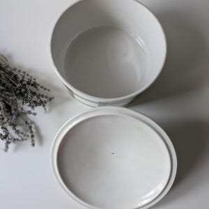 May include: White ceramic container with a lid, featuring a round shape. The container has a light green and white striped design around the rim. Dried lavender sprigs are visible on the left side, suggesting a storage or decorative theme.
