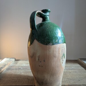 May include: A tall, antique stoneware jug with a green glazed top and handle. The lower portion of the jug is a natural, unglazed tan color. The jug sits on a rustic wooden surface.