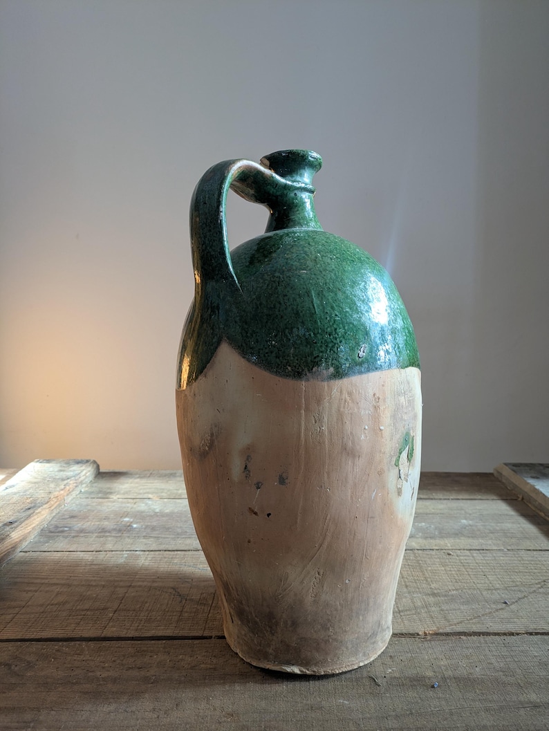 May include: A tall, antique stoneware jug with a green glazed top and handle. The lower portion of the jug is a natural, unglazed tan color. The jug sits on a rustic wooden surface.