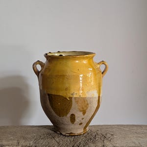 Może przedstawiać: Ceramiczne naczynie z dwoma bocznymi uchwytami, posiadające błyszczącą musztardowo-żółtą glazurę w górnej części oraz matowe, nieszkliwione wykończenie z beżowej gliny w dolnej części, z widocznymi zaciekami i różnicami w nałożeniu glazury.