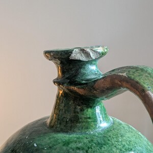 May include: Close-up of a green glazed ceramic jug with a handle. The jug has a rounded body and a narrow neck with a chipped rim. The handle is brown and green. The glaze has a crackled appearance.
