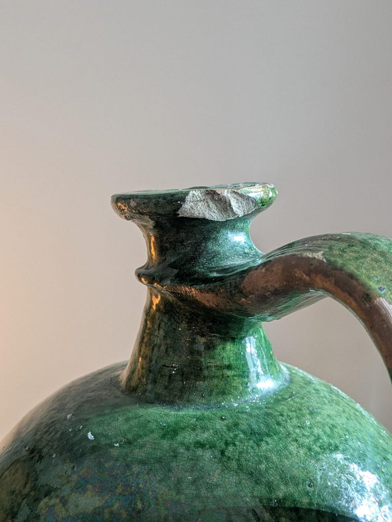 May include: Close-up of a green glazed ceramic jug with a handle. The jug has a rounded body and a narrow neck with a chipped rim. The handle is brown and green. The glaze has a crackled appearance.