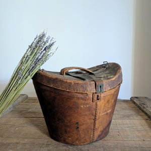 Pu&ograve; includere: Una cappelliera in pelle marrone usurata con coperchio curvo, manico superiore e chiusura a scatto in metallo. La scatola presenta cuciture visibili lungo i lati e una consistenza invecchiata. Un mazzo di lavanda essiccata &egrave; posizionato accanto.