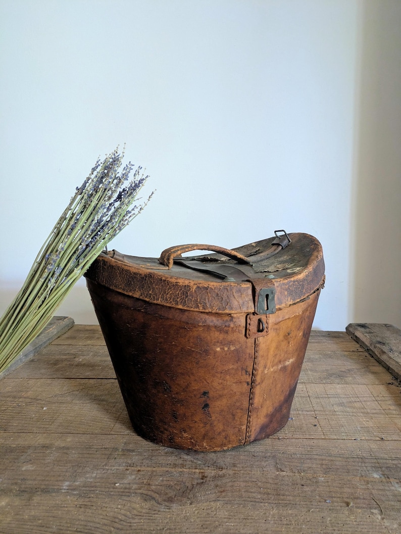 Pu&ograve; includere: Una cappelliera in pelle marrone usurata con coperchio curvo, manico superiore e chiusura a scatto in metallo. La scatola presenta cuciture visibili lungo i lati e una consistenza invecchiata. Un mazzo di lavanda essiccata &egrave; posizionato accanto.