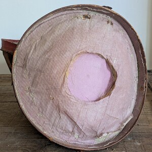 Pu&ograve; includere: Una cappelliera rotonda in stile vintage con bordo esterno marrone e rivestimento interno in tessuto a righe sottili rosa chiaro e bianco. Il centro della base interna presenta un ritaglio circolare che rivela un materiale solido in schiuma viola chiaro.