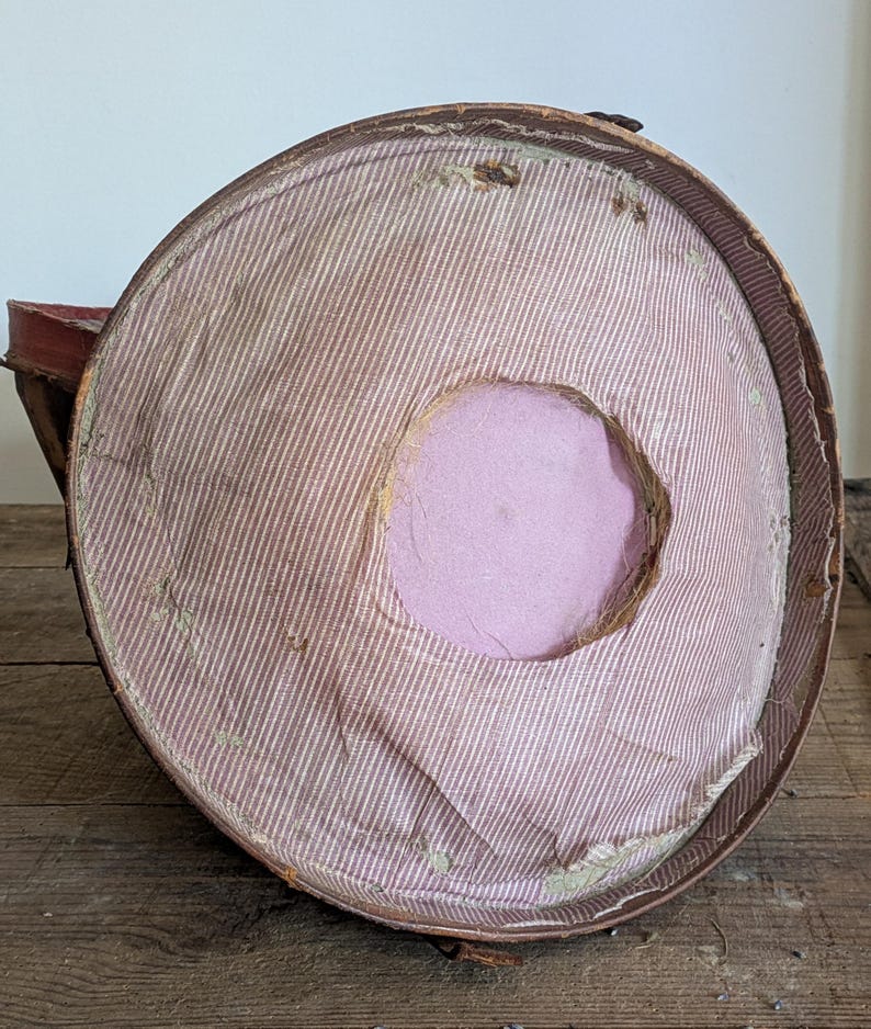 Pu&ograve; includere: Una cappelliera rotonda in stile vintage con bordo esterno marrone e rivestimento interno in tessuto a righe sottili rosa chiaro e bianco. Il centro della base interna presenta un ritaglio circolare che rivela un materiale solido in schiuma viola chiaro.