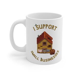 May include: White ceramic mug with a wraparound design. The design features a cartoon illustration of a wooden house with the text "I Support Small Businesses". The house has a red roof and a sign that says "Hats".