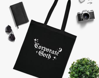 Puede incluir: Bolso tote negro con la frase "Corporate Goth" en letras góticas blancas. El bolso tiene dos asas negras. También hay un cuaderno negro, gafas de sol, una cámara y una pequeña planta.