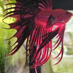 Może przedstawiać: Ozdoba w kształcie rybki betta w kolorze różowym i czarnym z błyszczącym wykończeniem. Ozdoba jest zawieszona na czarnym metalowym stojaku.