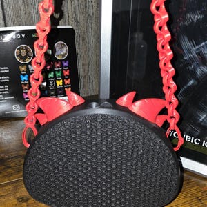 May include: A black, textured, oval-shaped bag with a red chain strap and red decorative accents. The bag has a unique design and is suspended by a chain. The bag is on a wooden surface.