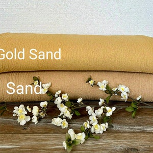Puede incluir: Dos piezas de tela, una de color marrón claro etiquetada como "Sand" y la otra de color marrón oscuro etiquetada como "Gold Sand". La tela es texturizada y tiene un aspecto arrugado. Una ramita de flores blancas está delante de la tela.