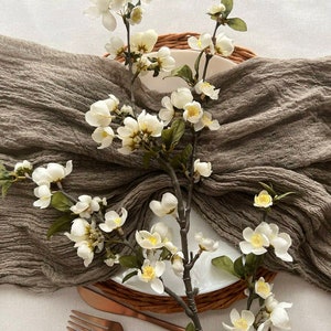 May include: A brown linen table runner with white blossoms and green leaves. The runner is draped over a woven placemat with a white plate.