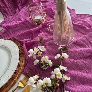 May include: A purple table runner with a white flower sprig, a gold candle holder, and a clear glass vase with dried flowers.