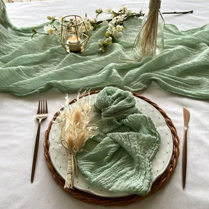 Puede incluir: Un camino de mesa verde salvia con textura tejida, un plato blanco con una servilleta verde salvia, un arreglo de flores secas y candelabros de metal dorado con velas.