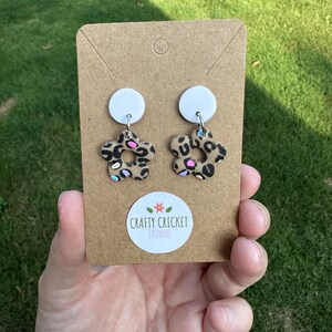 May include: A pair of leopard print flower earrings with colourful accents. The earrings are displayed on a brown card with the text "Crafty Cricket Studio".