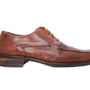 Puede incluir: Un zapato de vestir individual de cuero marrón con punta cuadrada y cierre de cordones. El zapato tiene un aspecto desgastado y envejecido, con costuras visibles y una suela oscura. El tacón mide aproximadamente 2,5 cm de alto.