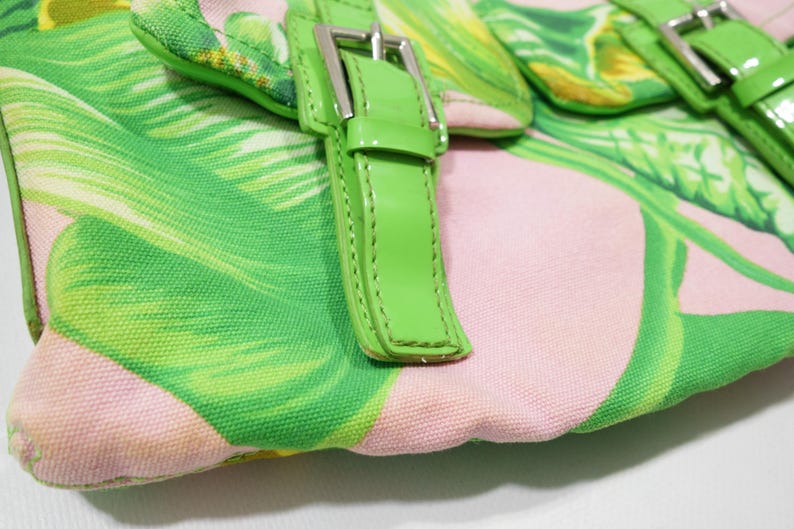K&ouml;nnte beinhalten: Nahaufnahme einer Handtasche mit tropischem Bl&auml;ttermuster in Gr&uuml;n und Rosa. Die Tasche hat leuchtend gr&uuml;ne Lederriemen und silberne Schnallen. Der Stoff hat eine leinwandartige Textur.