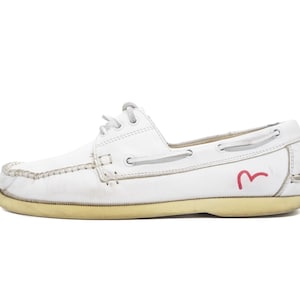 May include: White leather boat shoe with a light tan sole. The shoe features white laces, stitching details, and a red logo on the side. The shoe is designed for casual wear and has a classic, timeless style.