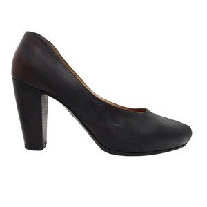 May include: A dark brown leather pump shoe with a rounded toe and a thick, sturdy block heel. The shoe features a smooth, matte finish across the upper and a slightly lighter brown tone on the heel, showcasing a classic, simple silhouette.