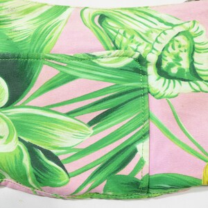 K&ouml;nnte beinhalten: Eine rosa Stofftasche mit einem lebendigen gr&uuml;nen Blumenmuster. Das Design zeigt gro&szlig;e, stilisierte gr&uuml;ne Bl&auml;tter und Bl&uuml;ten. Die Tasche hat eine Tasche auf der Vorderseite und ist wahrscheinlich eine Clutch oder eine kleine Handtasche.