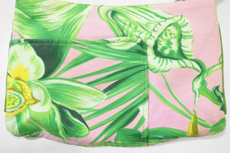 K&ouml;nnte beinhalten: Eine rosa Stofftasche mit einem lebendigen gr&uuml;nen Blumenmuster. Das Design zeigt gro&szlig;e, stilisierte gr&uuml;ne Bl&auml;tter und Bl&uuml;ten. Die Tasche hat eine Tasche auf der Vorderseite und ist wahrscheinlich eine Clutch oder eine kleine Handtasche.
