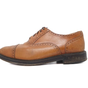 Peut inclure: Une chaussure oxford en cuir beige dotée d'un bout droit avec des perforations décoratives de type brogue le long des coutures et des œillets. La chaussure possède une semelle et un talon en caoutchouc marron foncé avec des lacets fins.