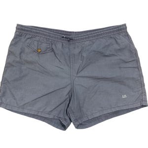 May include: A pair of slate blue swim shorts with an elastic waistband and drawstring. The shorts feature a single pocket with a button closure and a small logo near the hem. The fabric has a slightly textured appearance.