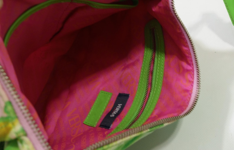 K&ouml;nnte beinhalten: Innenansicht einer Versus Handtasche. Die Tasche hat ein leuchtend pinkfarbenes Innenfutter mit einem gr&uuml;nen Rei&szlig;verschluss und Zierleisten. Die Au&szlig;enseite hat ein Blumenmuster. Ein kleines Etikett mit dem Wort "VERSUS" ist im Inneren sichtbar.
