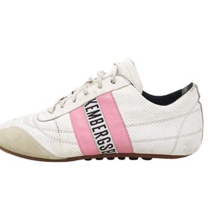 May include: A white perforated leather sneaker with a beige suede toe cap and a pink vertical stripe featuring the black text BIKEMBERGS. The shoe includes white laces, a dark interior lining, and a brown rubber sole with a textured tread pattern.