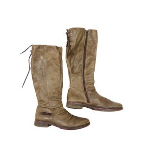 May include: A pair of knee-high, olive-green leather boots. One boot has a side zipper, while the other has a lace-up back. Both boots have a low heel and a distressed finish, suggesting a vintage style.
