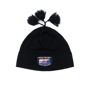 Puede incluir: Gorro de punto negro con tres borlas negras. El gorro presenta un parche rectangular con el texto "POLO SPORT RALPH LAUREN" y "PSRL67 ARCTIC CHALLENGE" en blanco, rojo y azul.