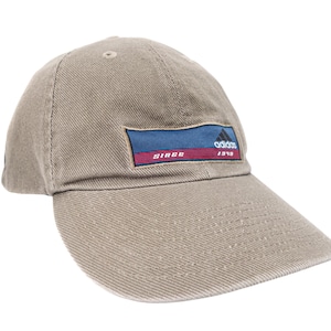 May include: A tan baseball cap featuring a rectangular patch on the front. The patch has a blue background with a black Adidas logo, a red horizontal stripe, and the white text "SINCE 1949". The cap has a curved brim and stitched detailing.