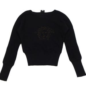 May include: Black knitted sweater with long sleeves and a round neck. The sweater has a Medusa head emblem in the centre. The sleeves are slightly puffed at the shoulder and taper to the wrist. The material is a ribbed knit.