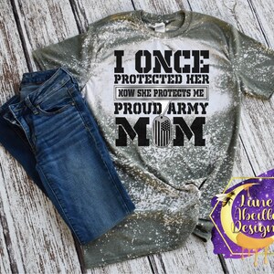 Puede incluir: Una camiseta verde y blanca blanqueada con el texto "I once protected her now she protects me proud army mom" en texto negro con una placa de identificaci&oacute;n plateada con una bandera estadounidense.