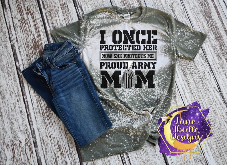 Puede incluir: Una camiseta verde y blanca blanqueada con el texto "I once protected her now she protects me proud army mom" en texto negro con una placa de identificaci&oacute;n plateada con una bandera estadounidense.